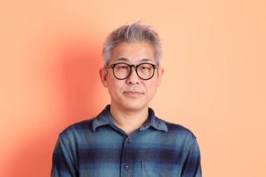 Yeşil ekose giysili Asyalı son sınıf öğrencisi gözlüklü, ayakta duran ve turuncu arka planda sırıtan. Turist, akıllı gündelik.