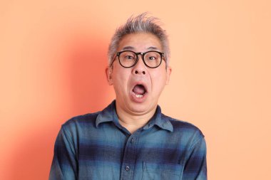 Yeşil ekose giysili Asyalı son sınıf öğrencisi gözlüklü, sürpriz jestli ve turuncu arka planda etkileyici izole edilmiş. Turist, akıllı gündelik.