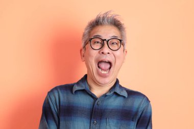 Yeşil ekose giysili Asyalı son sınıf öğrencisi gözlüklü, sürpriz jestli ve turuncu arka planda etkileyici izole edilmiş. Turist, akıllı gündelik.