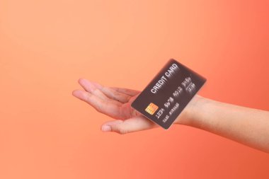 Yeşil ekose elbiseli Asyalı son sınıf öğrencisi gözlük takıyor ve turuncu arka planda kredi kartı taşıyor. Turist, akıllı gündelik.