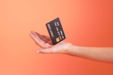 Yeşil ekose elbiseli Asyalı son sınıf öğrencisi gözlük takıyor ve turuncu arka planda kredi kartı taşıyor. Turist, akıllı gündelik.