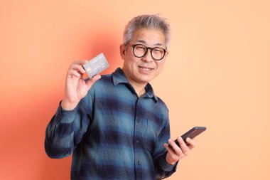 Yeşil ekose elbiseli Asyalı son sınıf öğrencisi. Gözlük takıyor. Kredi kartı taşıyor ve turuncu arka planda cep telefonu var. Turist, akıllı gündelik.