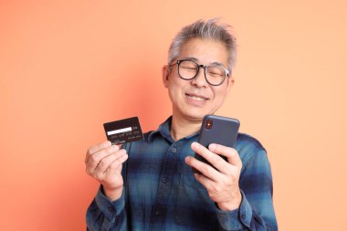 Yeşil ekose elbiseli Asyalı son sınıf öğrencisi. Gözlük takıyor. Kredi kartı taşıyor ve turuncu arka planda cep telefonu var. Turist, akıllı gündelik.