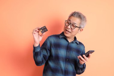 Yeşil ekose elbiseli Asyalı son sınıf öğrencisi. Gözlük takıyor. Kredi kartı taşıyor ve turuncu arka planda cep telefonu var. Turist, akıllı gündelik.