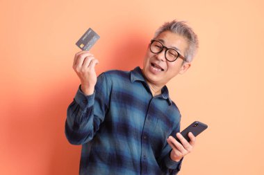 Yeşil ekose elbiseli Asyalı son sınıf öğrencisi. Gözlük takıyor. Kredi kartı taşıyor ve turuncu arka planda cep telefonu var. Turist, akıllı gündelik.