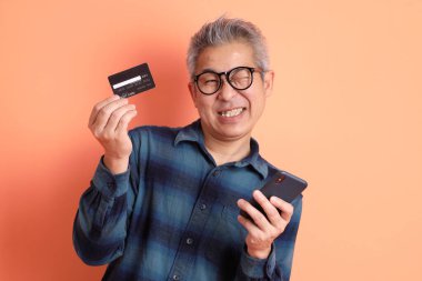 Yeşil ekose elbiseli Asyalı son sınıf öğrencisi. Gözlük takıyor. Kredi kartı taşıyor ve turuncu arka planda cep telefonu var. Turist, akıllı gündelik.