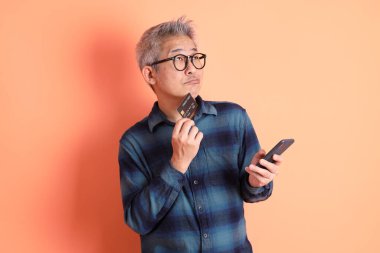 Yeşil ekose elbiseli Asyalı son sınıf öğrencisi. Gözlük takıyor. Kredi kartı taşıyor ve turuncu arka planda cep telefonu var. Turist, akıllı gündelik.