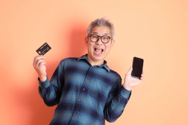 Yeşil ekose elbiseli Asyalı son sınıf öğrencisi. Gözlük takıyor. Kredi kartı taşıyor ve turuncu arka planda cep telefonu var. Turist, akıllı gündelik.