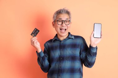 Yeşil ekose elbiseli Asyalı son sınıf öğrencisi. Gözlük takıyor. Kredi kartı taşıyor ve turuncu arka planda cep telefonu var. Turist, akıllı gündelik.