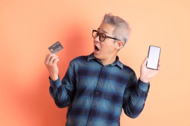 Yeşil ekose elbiseli Asyalı son sınıf öğrencisi. Gözlük takıyor. Kredi kartı taşıyor ve turuncu arka planda cep telefonu var. Turist, akıllı gündelik.