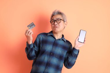 Yeşil ekose elbiseli Asyalı son sınıf öğrencisi. Gözlük takıyor. Kredi kartı taşıyor ve turuncu arka planda cep telefonu var. Turist, akıllı gündelik.