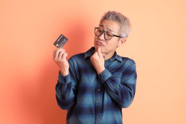 Yeşil ekose elbiseli Asyalı son sınıf öğrencisi gözlük takıyor ve turuncu arka planda kredi kartı taşıyor. Turist, akıllı gündelik.