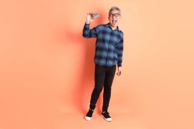 Yeşil ekose elbiseli Asyalı son sınıf öğrencisi gözlük takıyor ve turuncu arka planda kredi kartı taşıyor. Turist, akıllı gündelik.