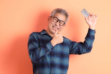 Yeşil ekose elbiseli Asyalı son sınıf öğrencisi gözlük takıyor ve turuncu arka planda kredi kartı taşıyor. Turist, akıllı gündelik.