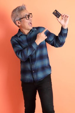 Yeşil ekose elbiseli Asyalı son sınıf öğrencisi gözlük takıyor ve turuncu arka planda kredi kartı taşıyor. Turist, akıllı gündelik.