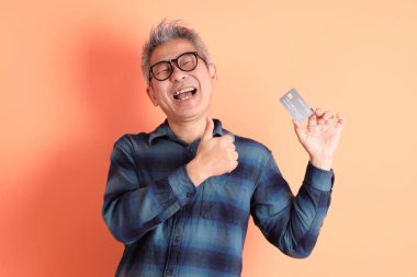 Yeşil ekose elbiseli Asyalı son sınıf öğrencisi gözlük takıyor ve turuncu arka planda kredi kartı taşıyor. Turist, akıllı gündelik.
