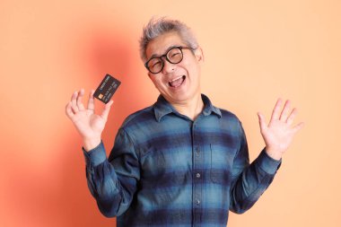 Yeşil ekose elbiseli Asyalı son sınıf öğrencisi gözlük takıyor ve turuncu arka planda kredi kartı taşıyor. Turist, akıllı gündelik.