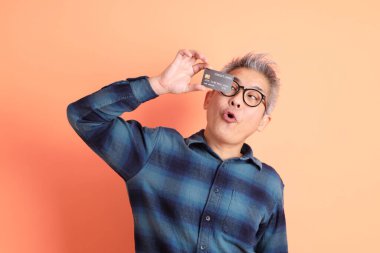 Yeşil ekose elbiseli Asyalı son sınıf öğrencisi gözlük takıyor ve turuncu arka planda kredi kartı taşıyor. Turist, akıllı gündelik.