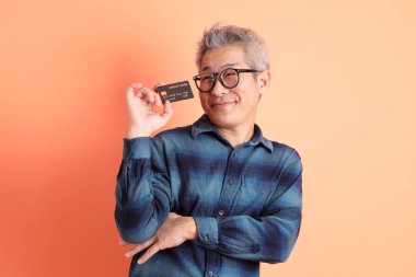 Yeşil ekose elbiseli Asyalı son sınıf öğrencisi gözlük takıyor ve turuncu arka planda kredi kartı taşıyor. Turist, akıllı gündelik.