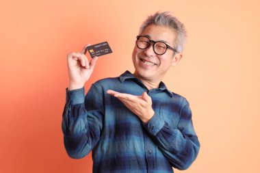 Yeşil ekose elbiseli Asyalı son sınıf öğrencisi gözlük takıyor ve turuncu arka planda kredi kartı taşıyor. Turist, akıllı gündelik.