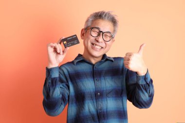 Yeşil ekose elbiseli Asyalı son sınıf öğrencisi gözlük takıyor ve turuncu arka planda kredi kartı taşıyor. Turist, akıllı gündelik.