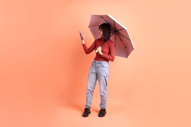 Kırmızı uzun kollu tişörtlü kısa saçlı Asyalı kadın. El ele tutuşurken mavi ve pembe şemsiyeli jest turuncu arka planda izole edilmiş. Gen Z
