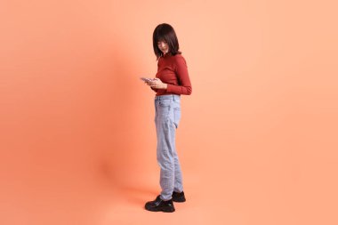 Kırmızı, uzun kollu tişörtlü kısa saçlı Asyalı kadın. Turuncu arka planda cep telefonu bulundurduğunu gösteren bir jest. Gen Z