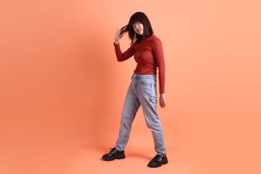 Kırmızı uzun kollu tişörtlü kısa saçlı Asyalı kadın. Turuncu arka planda duran ve gülümseyen. Gen Z