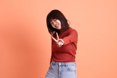 Kısa saçlı, kısa kollu, uzun kollu, kırmızı tişörtlü, barış işareti olan, V işareti olan, turuncu arka planda iki parmağı olan Asyalı bir kadın. Gen Z