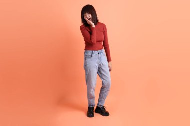 Kırmızı uzun kollu tişörtlü kısa saçlı Asyalı kadın. Turuncu arka planda duran ve gülümseyen. Gen Z