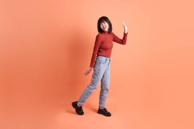 Kırmızı uzun kollu tişörtlü kısa saçlı Asyalı kadın. Turuncu arka planda duran ve gülümseyen. Gen Z