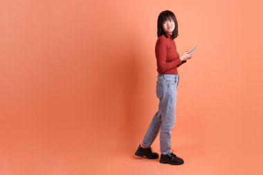 Kırmızı, uzun kollu tişörtlü kısa saçlı Asyalı kadın. Turuncu arka planda cep telefonu bulundurduğunu gösteren bir jest. Gen Z