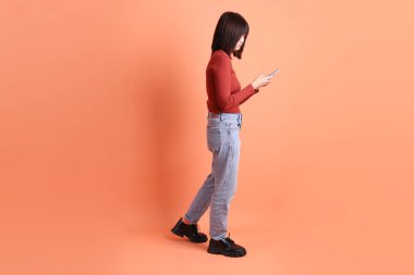 Kırmızı, uzun kollu tişörtlü kısa saçlı Asyalı kadın. Turuncu arka planda cep telefonu bulundurduğunu gösteren bir jest. Gen Z