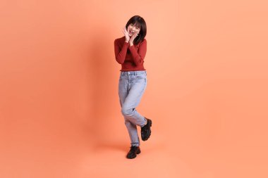 Kırmızı uzun kollu tişörtlü kısa saçlı Asyalı kadın. Bağırma hareketi yapıyor. Elinde turuncu arka planda izole edilmiş bir megafon tutuyor. Gen Z