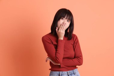 Kısa saçlı Asyalı kadın, kırmızı uzun kollu tişörtlü, ağzı kapalı, turuncu arka planda izole edilmiş sessiz bir jest. Gen Z