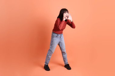 Kırmızı uzun kollu tişörtlü kısa saçlı Asyalı kadın. Bağırma hareketi yapıyor. Elinde turuncu arka planda izole edilmiş bir megafon tutuyor. Gen Z