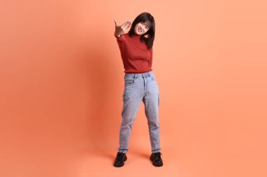 Kırmızı, uzun kollu tişörtlü kısa saçlı Asyalı kadın. Buraya gelip turuncu arka planda izole bir şekilde el sallıyor. Gen Z