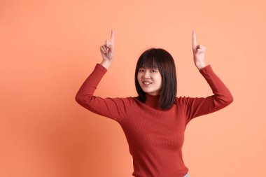 Kırmızı uzun kollu tişörtlü kısa saçlı Asyalı kadın turuncu arka planda izole edilmiş bir işaret hareketi yapıyor. Gen Z