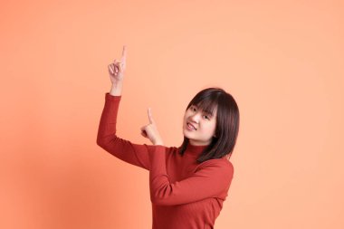 Kırmızı uzun kollu tişörtlü kısa saçlı Asyalı kadın turuncu arka planda izole edilmiş bir işaret hareketi yapıyor. Gen Z