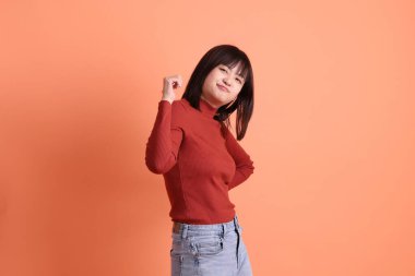 Kırmızı uzun kollu tişörtlü kısa saçlı Asyalı kadın turuncu arka planda izole edilmiş bir işaret hareketi yapıyor. Gen Z
