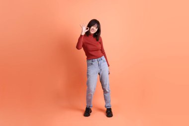 Kırmızı uzun kollu tişörtlü kısa saçlı Asyalı kadın başparmak hareketi yapıyor ya da turuncu arka planda izole olduğunu onaylıyor. Gen Z