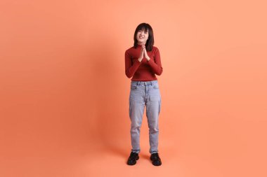 Kırmızı uzun kollu tişörtlü kısa saçlı Asyalı kadın turuncu arka planda izole edilmiş selamlama jesti yapıyor. Gen Z