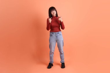Kırmızı uzun kollu tişörtlü kısa saçlı Asyalı kadın turuncu arka planda izole edilmiş bir işaret hareketi yapıyor. Gen Z