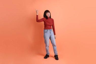Kırmızı uzun kollu tişörtlü kısa saçlı Asyalı kadın turuncu arka planda izole edilmiş bir işaret hareketi yapıyor. Gen Z