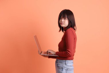 Kırmızı, uzun kollu tişörtlü kısa saçlı Asyalı kadın. Dizüstü bilgisayarını turuncu arka planda tutuyormuş. Gen Z