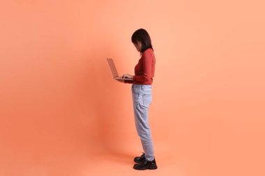 Kırmızı, uzun kollu tişörtlü kısa saçlı Asyalı kadın. Dizüstü bilgisayarını turuncu arka planda tutuyormuş. Gen Z