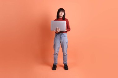 Kırmızı, uzun kollu tişörtlü kısa saçlı Asyalı kadın. Dizüstü bilgisayarını turuncu arka planda tutuyormuş. Gen Z