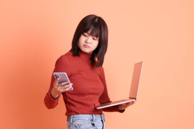 Kırmızı, uzun kollu tişörtlü kısa saçlı Asyalı kadın. Dizüstü bilgisayarını turuncu arka planda tutuyormuş. Gen Z