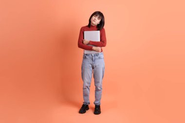 Kırmızı, uzun kollu tişörtlü kısa saçlı Asyalı kadın. Dizüstü bilgisayarını turuncu arka planda tutuyormuş. Gen Z