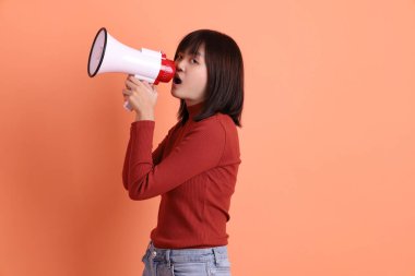 Kırmızı uzun kollu tişörtlü kısa saçlı Asyalı kadın. Bağırma hareketi yapıyor. Elinde turuncu arka planda izole edilmiş bir megafon tutuyor. Gen Z